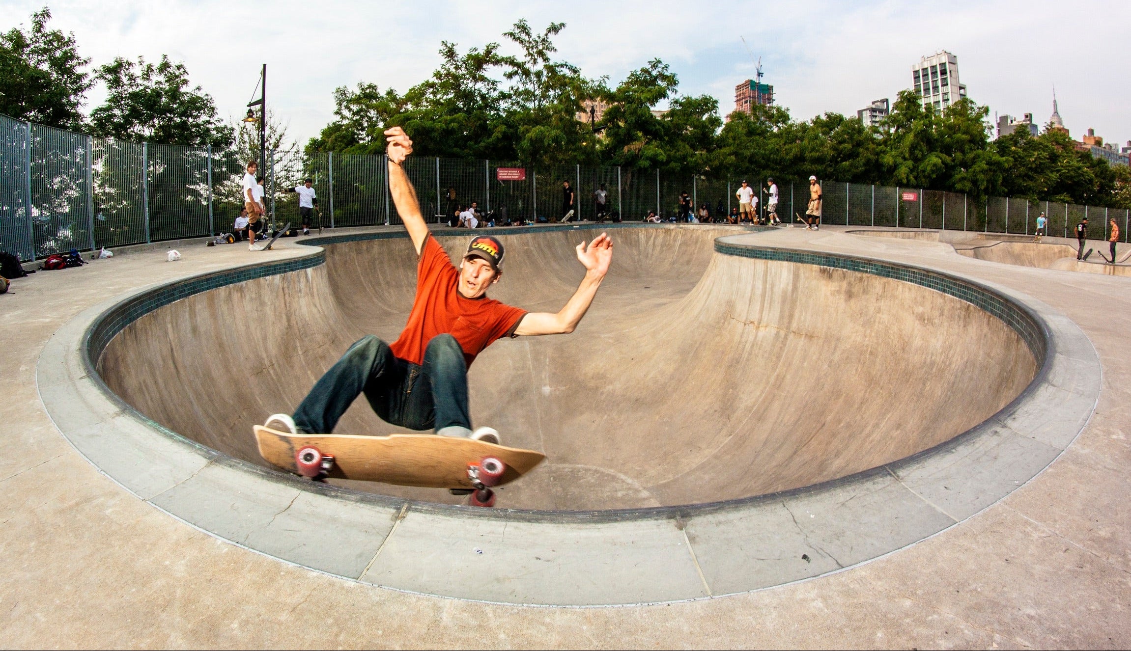 Person skateboarding in a concrete bowl at a skate park with trees and buildings in the background.